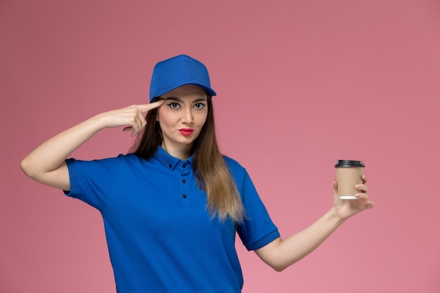 Front view female courier in blue uniform and cape holding delivery coffee cup on the pink wall job worker