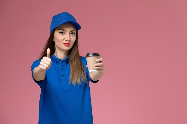 Front view female courier in blue uniform and cape holding delivery coffee cup posing on pink wall job
