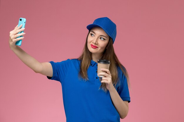 Front view female courier in blue uniform and cape holding delivery coffee cup taking selfie on pink wall 