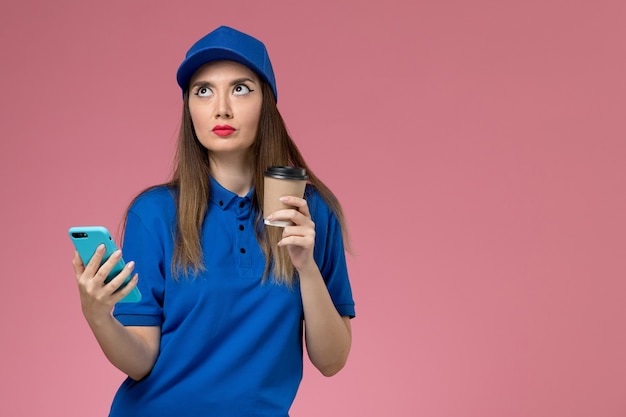 Front view female courier in blue uniform and cape holding delivery coffee cup and using phone on the pink desk 