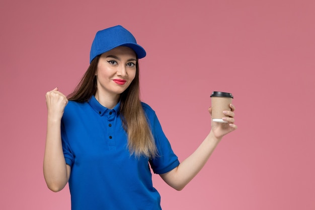 Front view female courier in blue uniform and cape holding delivery coffee cup with delighted expression on pink desk job worker woman