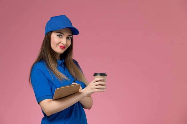 Front view female courier in blue uniform and cape holding delivery coffee cup writing and smiling on light-pink wall 
