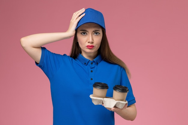 Женщина-курьер в синей форме и плаще, держащая кофейные чашки доставки и ее голову на розовой стене, вид спереди