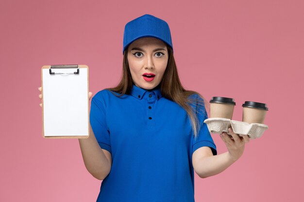 Front view female courier in blue uniform and cape holding delivery coffee cups and notepad on pink wall