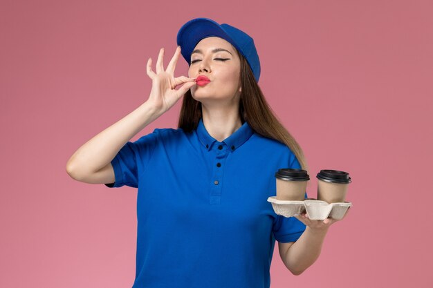 Front view female courier in blue uniform and cape holding delivery coffee cups on pink desk worker