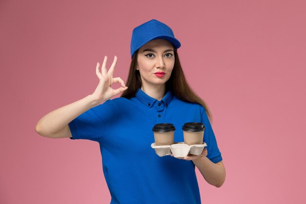 Front view female courier in blue uniform and cape holding delivery coffee cups on the pink desk