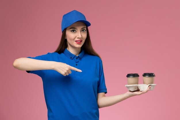 Front view female courier in blue uniform and cape holding delivery coffee cups on the pink wall job