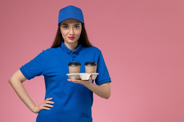 Front view female courier in blue uniform and cape holding delivery coffee cups on pink wall  worker female