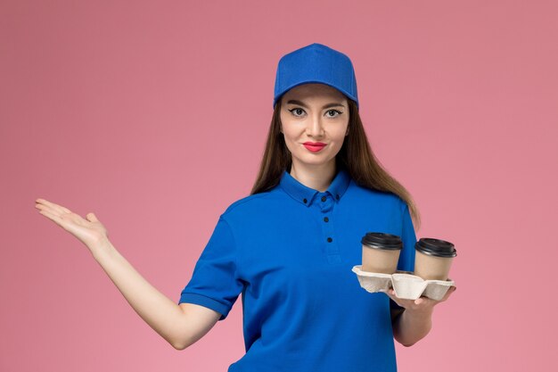 Front view female courier in blue uniform and cape holding delivery coffee cups on the pink wall