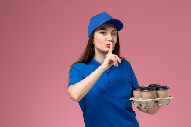 Front view female courier in blue uniform and cape holding delivery coffee cups and showing silence sign on pink wall 