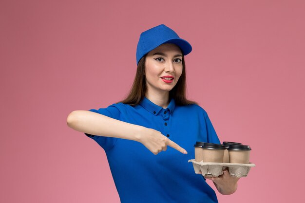 Женщина-курьер в синей униформе и накидке, держащая кофейные чашки с доставкой, улыбается на светло-розовой стене, вид спереди