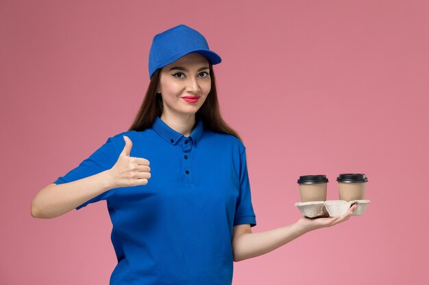 Женщина-курьер в синей форме и плаще, держащая кофейные чашки, улыбается на розовом столе, вид спереди