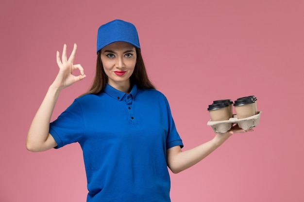 Front view female courier in blue uniform and cape holding delivery coffee cups and smiling on pink wall 