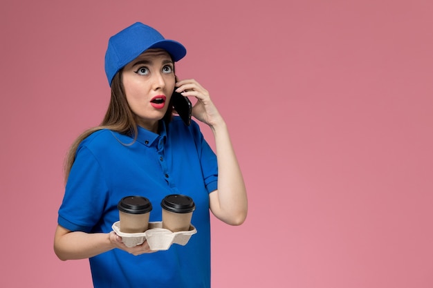 Front view female courier in blue uniform and cape holding delivery coffee cups talking on the phone on pink wall 