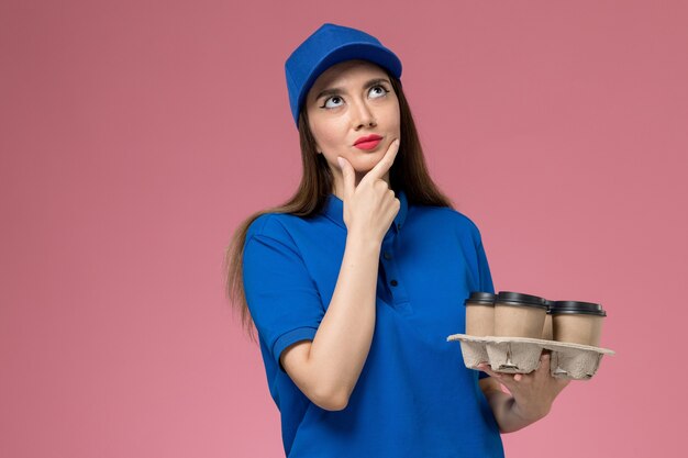 Front view female courier in blue uniform and cape holding delivery coffee cups and thinking on pink wall 