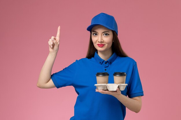 ピンクの壁に上げられた指で配達コーヒーカップを保持している青い制服と岬の正面図の女性の宅配便