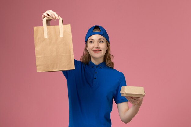 Front view female courier in blue uniform cape holding delivery packages on the pink wall, employee service delivery work