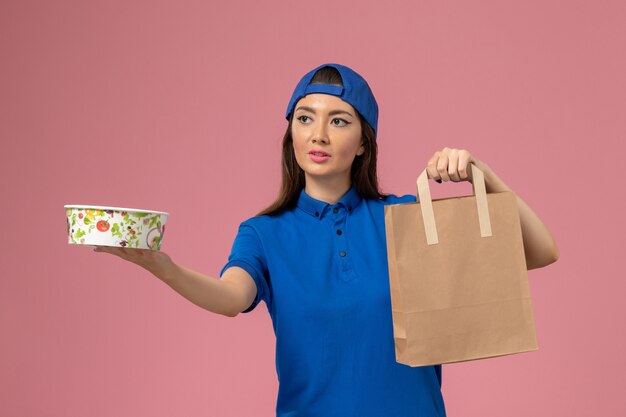 Front view female courier in blue uniform cape holding delivery paper package and bowl on pink wall, service employee delivering