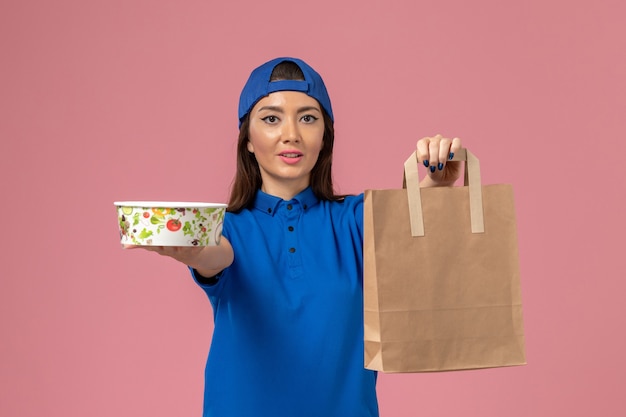 Front view female courier in blue uniform cape holding delivery paper package and bowl on pink wall, service employee job work delivering