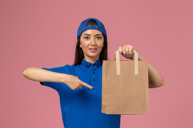 Женщина-курьер в синей форме, держащая бумажный пакет на розовой стене, вид спереди, доставляющий служащий