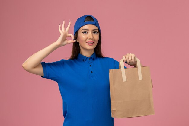 Front view female courier in blue uniform cape holding delivery paper package on the pink wall, work job service employee delivering