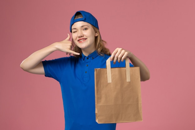 Front view female courier in blue uniform cape holding delivery paper package on pink wall, worker employee service delivery