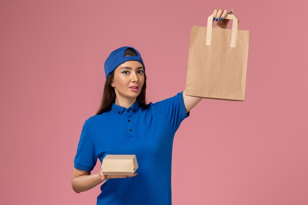Front view female courier in blue uniform cape holding different delivery packages on pink wall, service employee delivering work