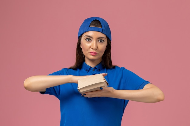 Женщина-курьер в синей униформе, держащая пустой маленький пакет для доставки на розовой стене, работник службы доставки компании, вид спереди