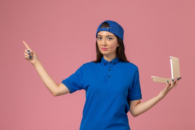 ピンクの壁に空の小さな配達パッケージを保持している青い制服ケープの正面図女性宅配便、従業員サービス会社の配達
