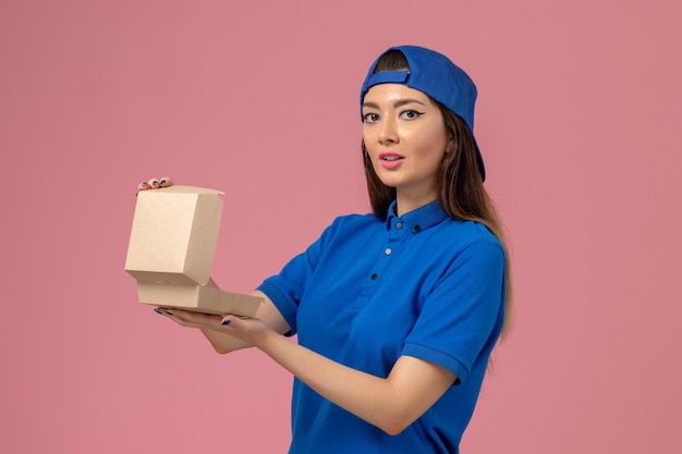 Женщина-курьер в синей униформе, держащая пустой маленький пакет для доставки на розовой стене, служба доставки работникам