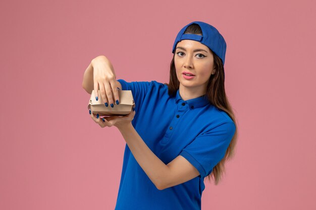 Front view female courier in blue uniform cape holding empty little delivery package on the pink wall, employee service job company delivery