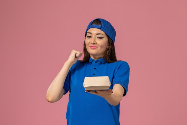 Front view female courier in blue uniform cape holding empty little delivery package and rejoicing on the pink wall, employee service company delivery