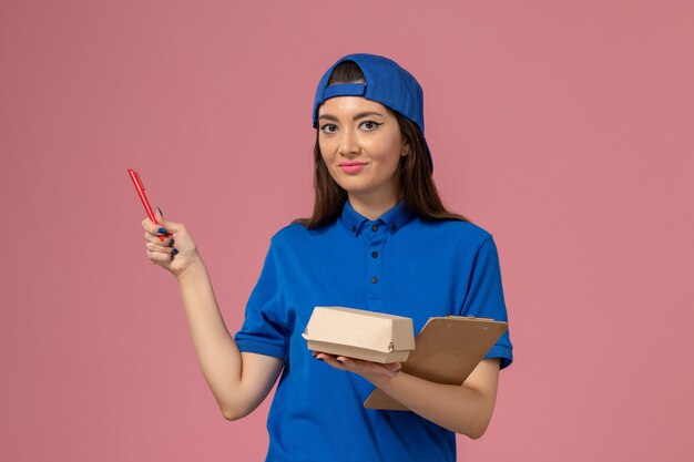 Front view female courier in blue uniform cape holding empty little delivery package with notepad on light pink wall, employee service delivery worker
