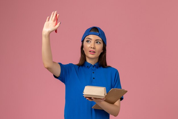 Front view female courier in blue uniform cape holding empty little delivery package with notepad on light pink wall, employee service work delivery