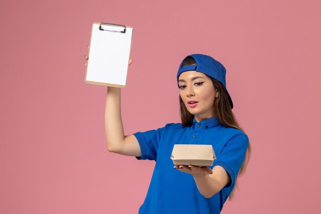 Front view female courier in blue uniform cape holding empty little delivery package with notepad on pink wall, employee service company delivery girl job
