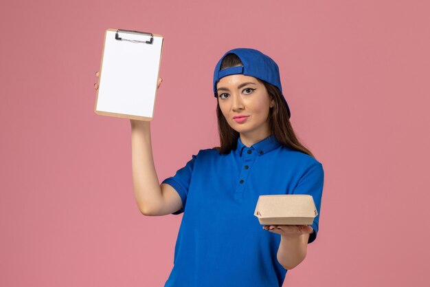 Front view female courier in blue uniform cape holding empty little delivery package with notepad on pink wall, employee service company delivery