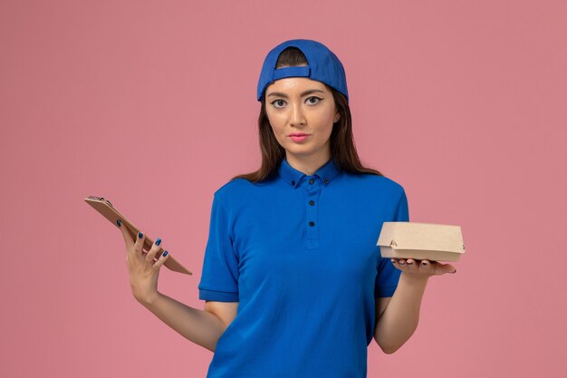 ピンクの壁にメモ帳付きの空の小さな配達パッケージ、従業員サービス会社の仕事の配達を保持している青い制服の岬の正面図の女性の宅配便