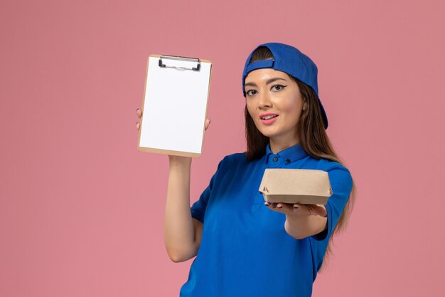 Front view female courier in blue uniform cape holding empty little delivery package with notepad on pink wall, employee service job company delivery worker