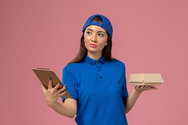 Женщина-курьер в синей униформе, вид спереди, держит пустой маленький пакет для доставки с блокнотом на розовой стене, служба доставки сотрудников компании