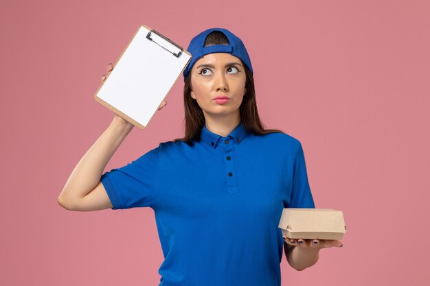 ピンクの壁にメモ帳付きの空の小さな配達パッケージ、従業員の仕事サービス会社の配達を保持している青い制服の岬の正面図の女性の宅配便