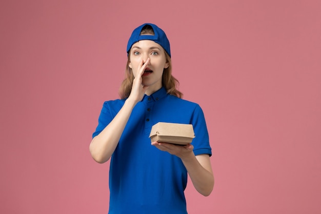 Front view female courier in blue uniform and cape holding little delivery food package calling out on the pink wall, delivery uniform service company