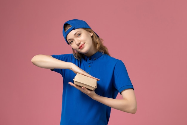 Free photo front view female courier in blue uniform and cape holding little delivery food package on light-pink wall, delivery job uniform service employee