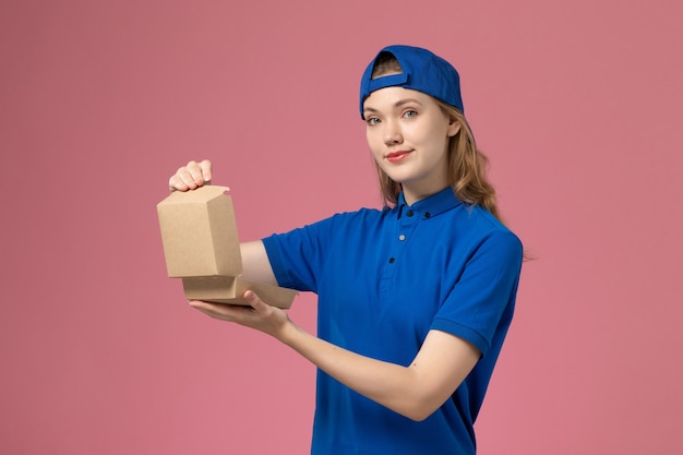 Front view female courier in blue uniform and cape holding little delivery food package on light-pink wall, delivery uniform service employee