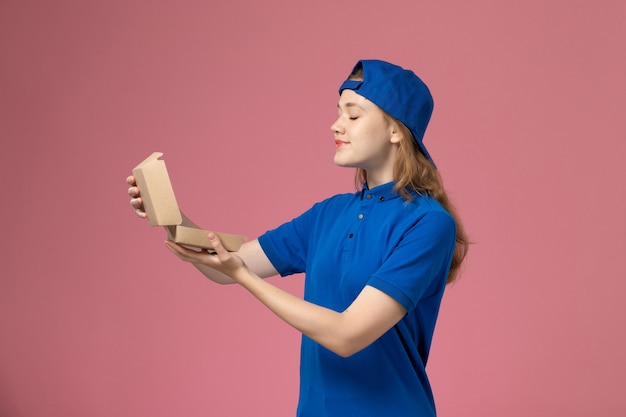 Front view female courier in blue uniform and cape holding little delivery food package on light-pink wall, delivery uniform service job employee