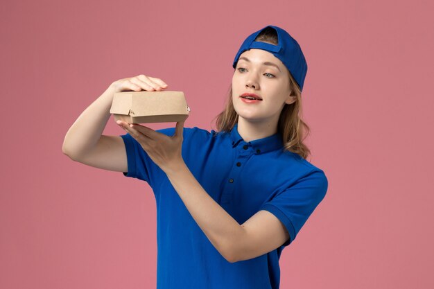 Front view female courier in blue uniform and cape holding little delivery food package on light-pink wall, work delivery uniform service employee