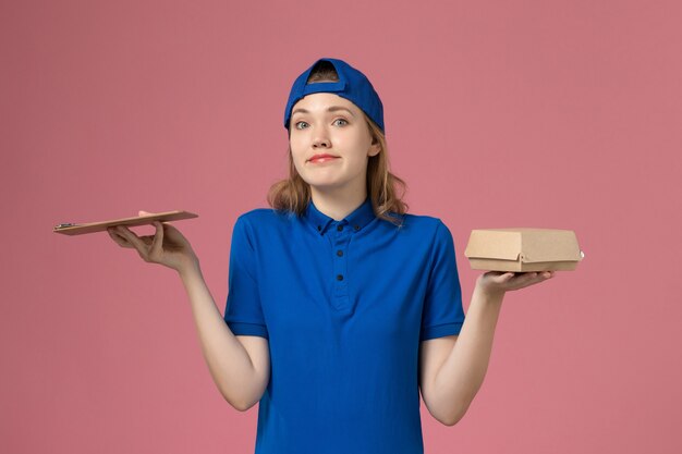 青いユニフォームとピンクの壁に小さな配達食品パッケージとメモ帳を保持している岬の正面図の女性の宅配便、配達サービスの従業員の仕事