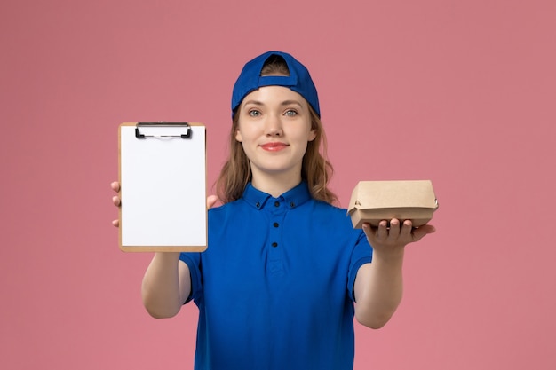 Front view female courier in blue uniform and cape holding little delivery food package and notepad on pink wall, delivery service employee