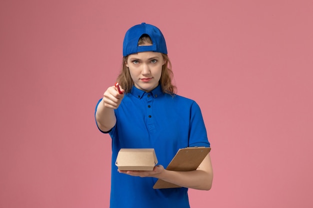 파란색 유니폼과 케이프 작은 배달 음식 패키지 메모장을 들고 분홍색 벽에 쓰는 전면보기 여성 택배, 배달 서비스 직원 소녀 작업