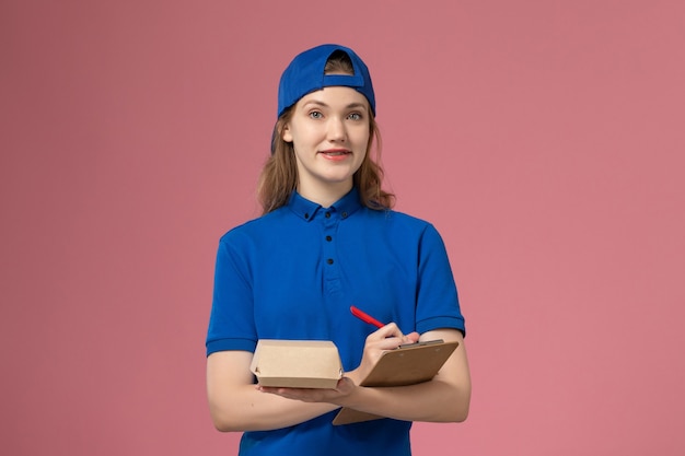 파란색 유니폼과 케이프 작은 배달 음식 패키지 메모장을 들고 분홍색 벽, 배달 서비스 직원 작업에 쓰는 전면보기 여성 택배