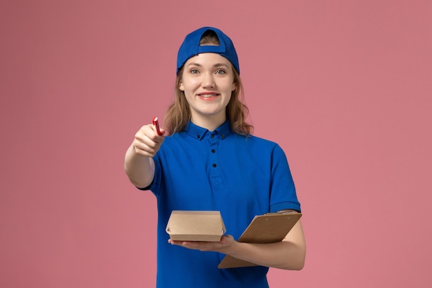 Front view female courier in blue uniform and cape holding little delivery food package notepad and writing on the pink wall, delivery service employee worker job